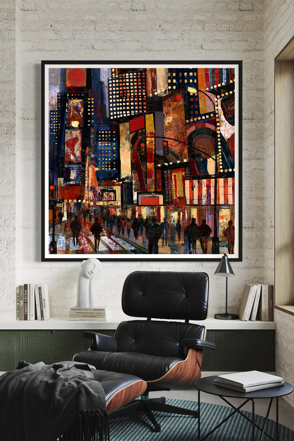 A large framed canvas art print depicting a vibrant, colorful Times Square at night, displayed on a textured light gray brick wall above a black leather lounge chair in a modern room setting.
