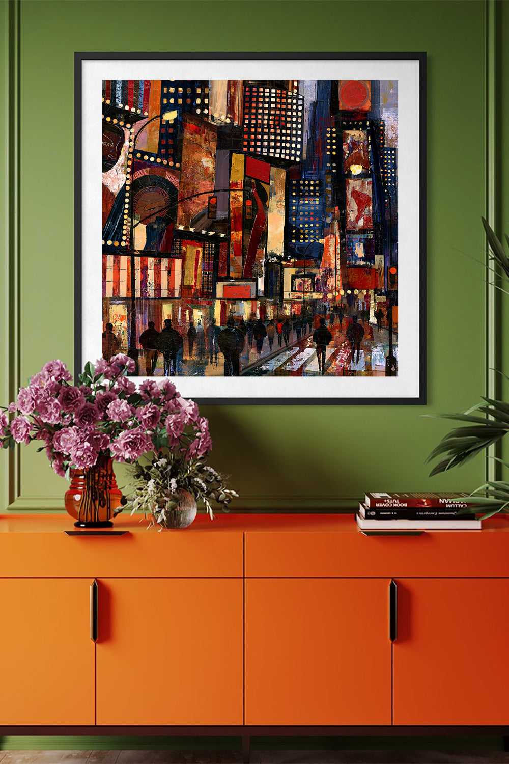 A framed art print depicting a vibrant, colorful Times Square city scene, displayed on a green wall above an orange sideboard with a vase of pink flowers and books.