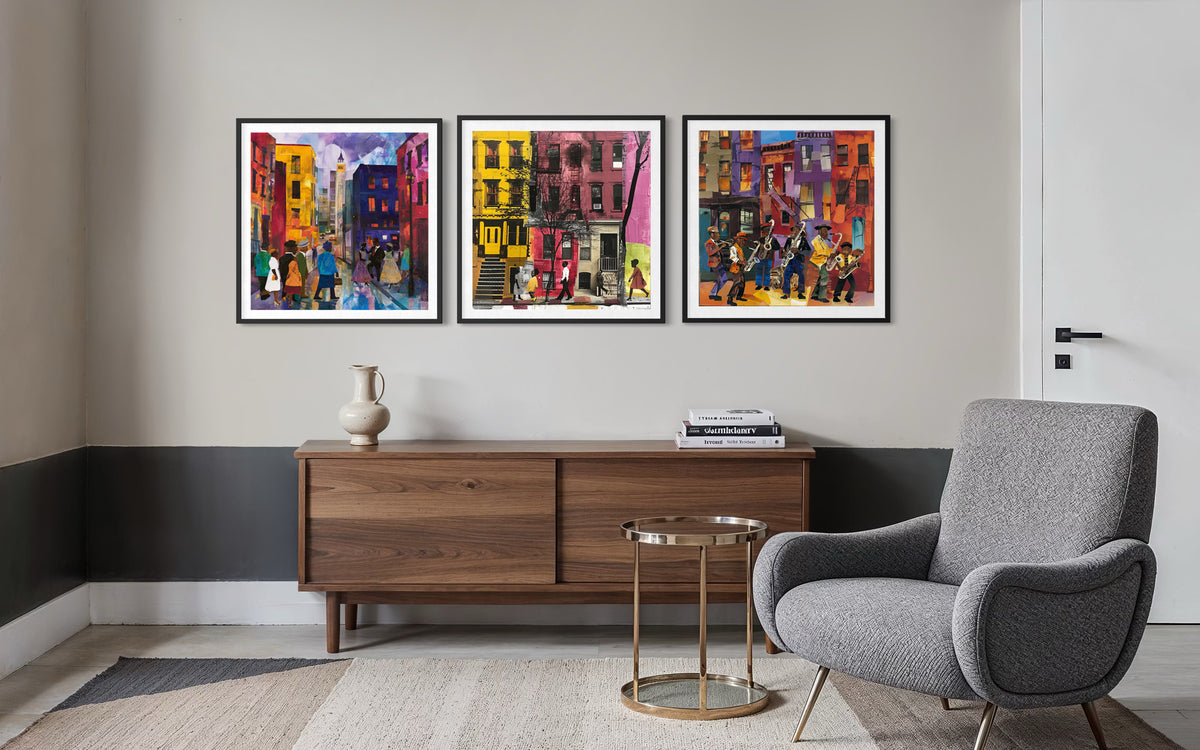 A styled interior room showing three framed colorful city street art prints hanging on a wall above a wooden sideboard, with a gray armchair and a small round side table in front.
