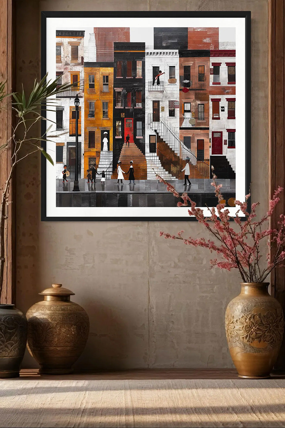 A framed art print depicting a stylized city street with colorful building facades and small figures, displayed on a wall above a shelf with decorative vases and branches.